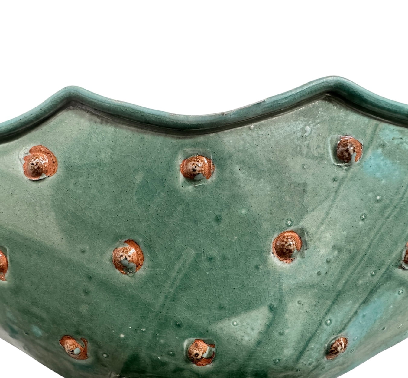 curved ceramic piece resembling a green pod, supported by a block plinth. The seafoam-green ceramic structure extends wide on either side, curving upward to form a wavy, boat-like shape. The outer surface is dotted with rows of small, raised, brown-orange, acorn-like protrusions. The top rim is layered with parallel, grooved ridges. The base is a vertical rectangular block with a heavily textured, bark-like surface in a dark green and black finish.