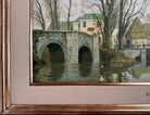 A naturalistic landscape oil painting by Michael Reilly titled "Mill Pond with Bridge" depicts a small town scene with a stone bridge over a mill pond, several buildings, and numerous bare trees under a grey sky