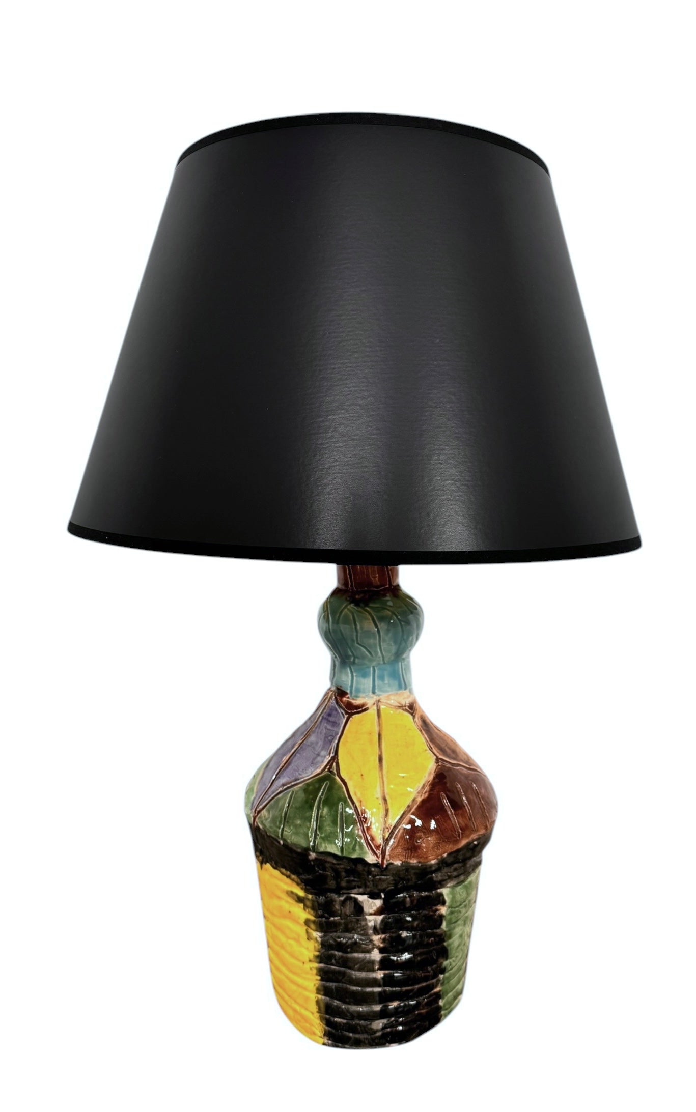 A hand-crafted ceramic table lamp featuring a vibrant, multi-colored base and a sleek black shade. The base is an upcycled artisan vase with a textured, hand-thrown appearance, decorated with geometric panels in shades of mustard yellow, forest green, cobalt blue, and burgundy, separated by thick black outlines. The lamp is topped with a minimalist, black tapered drum shade that provides a sharp contrast to the colorful, eclectic pottery below.
