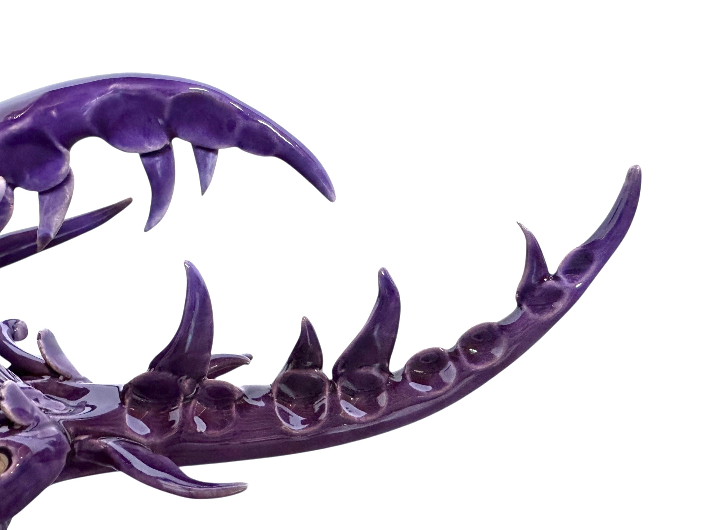 A glossy, deep purple ceramic sculpture of a beetle by Enza Fasano. The piece features a highly stylized and ornate design, blending insect-like anatomy with fantastical elements. It has a smooth, rounded shell and elongated, curved mandibles and appendages lined with sharp, thorn-like protrusions. The monochromatic glaze is reflective, highlighting the intricate sculptural details and textures of the avant-garde artwork.