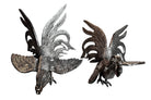 A pair of vintage, silver-plated brass fighting rooster figurines positioned in a confrontational stance against a white background. The rooster on the left is crouched low with its head down and tail feathers fanned high, while the rooster on the right stands more upright with its wings slightly flared. Both feature intricate feather detailing, a metallic patina showing hints of brass through the silver, and small, bright red glass eyes.