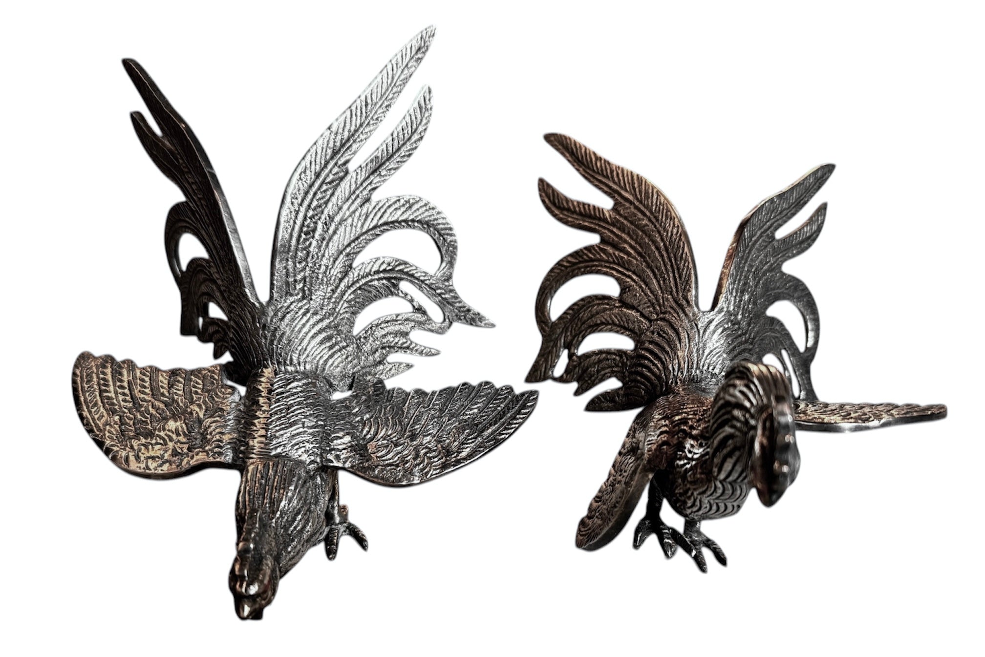 A pair of vintage, silver-plated brass fighting rooster figurines positioned in a confrontational stance against a white background. The rooster on the left is crouched low with its head down and tail feathers fanned high, while the rooster on the right stands more upright with its wings slightly flared. Both feature intricate feather detailing, a metallic patina showing hints of brass through the silver, and small, bright red glass eyes.