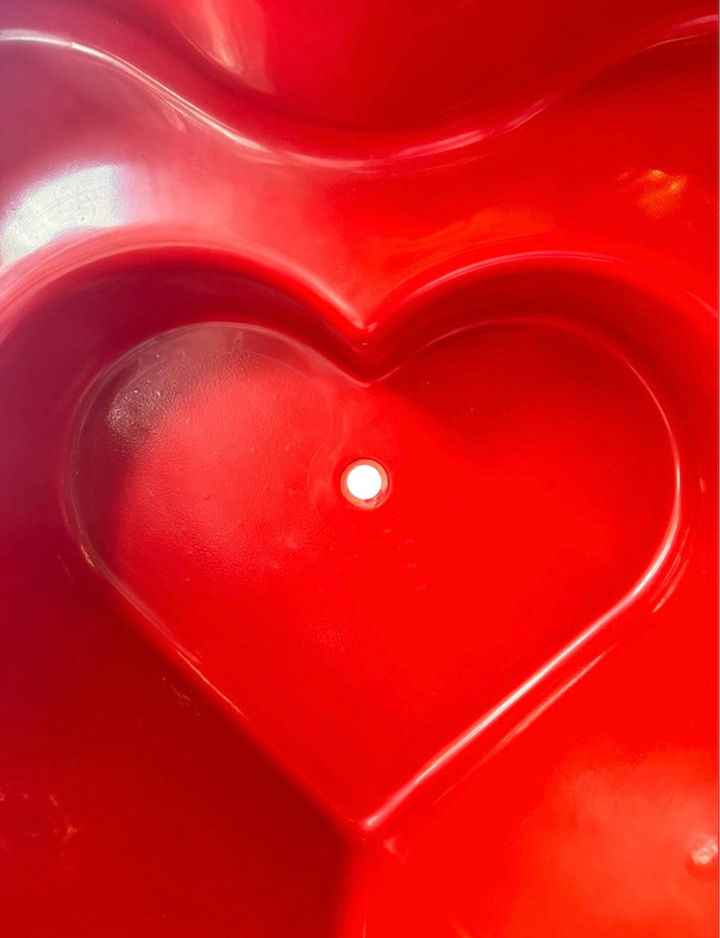 A top-down, straight-on view of a vintage, high-gloss cherry red bathtub. The tub basin is molded into a distinct double-tiered heart shape, with a shallower outer heart cascading into a deeper, smaller inner heart basin centered within a wide rectangular red deck.