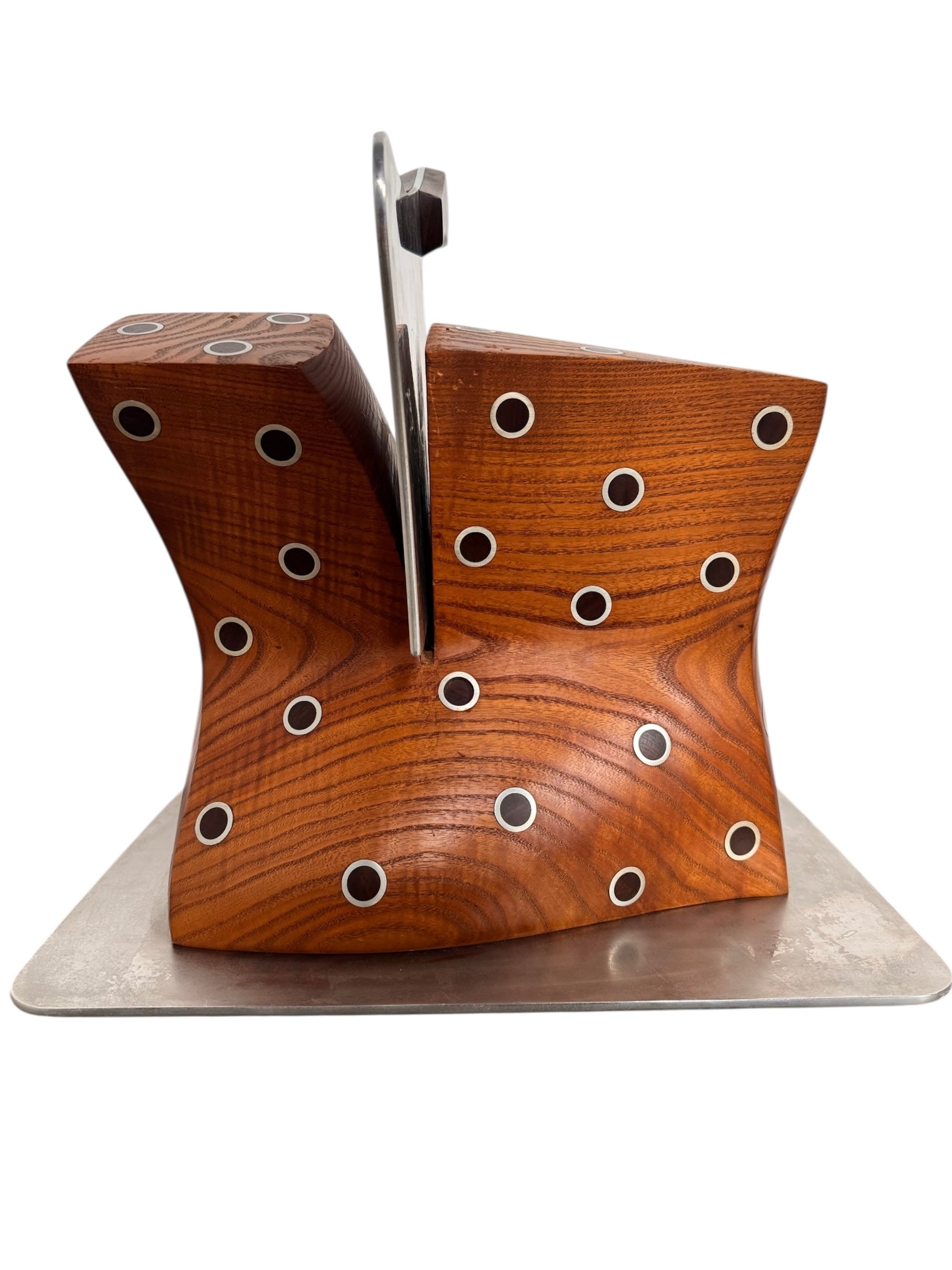 A wooden sculpture with a highly polished, reddish-brown grain, featuring two main asymmetrical, curved sections that converge at the top. Circular metal inlays with black centers are embedded across the surface of both wooden sections. A flat, polished metal blade-like element is inserted vertically between the two wooden sections, extending above the top surface. The sculpture rests on a metallic base.