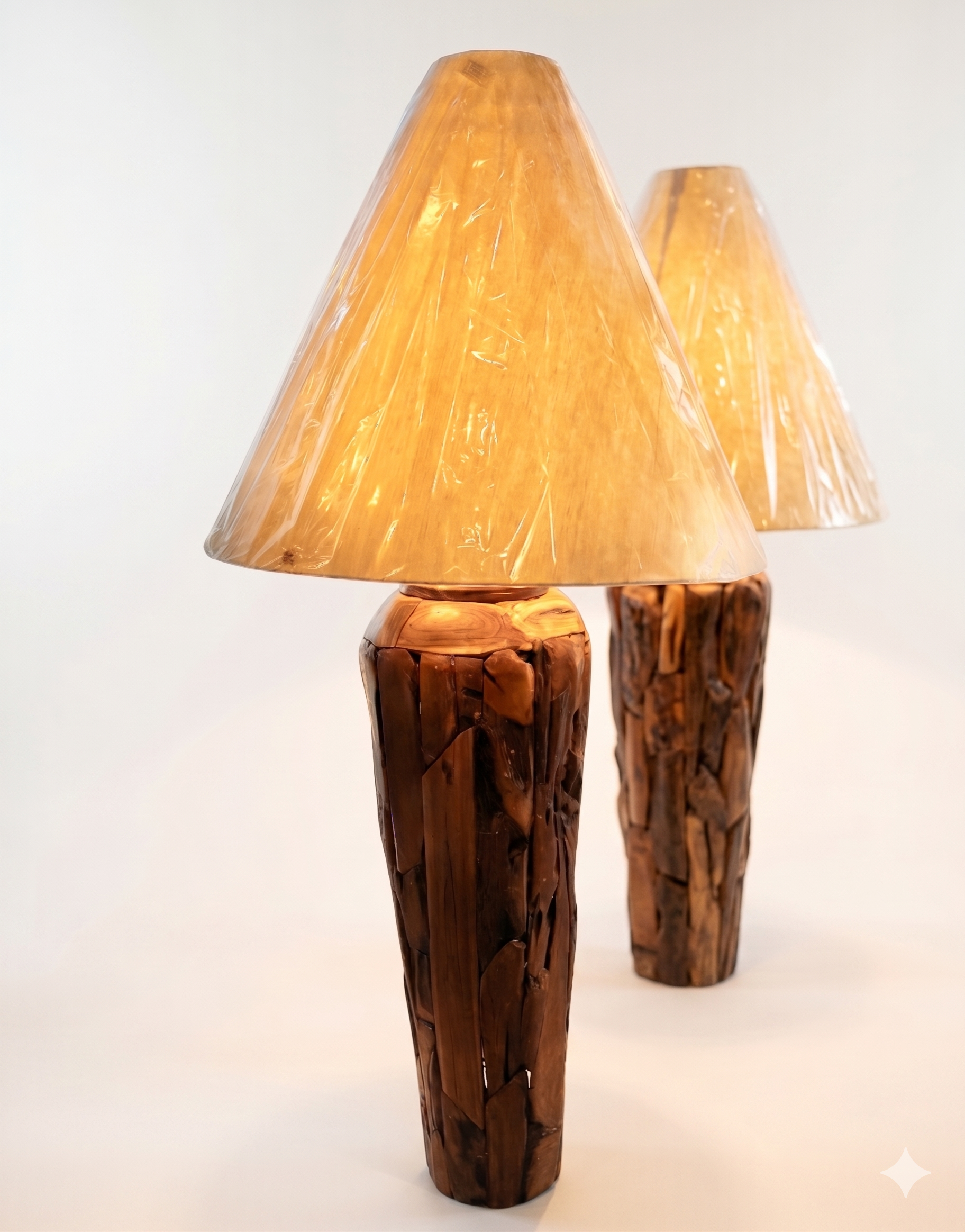 Two table lamps made from repurposed teak wood vases. The bases are tall and tapered, featuring a rugged, mosaic-like texture composed of various pieces of dark, polished teak wood with visible natural grain and crevices. Each lamp is topped with a large, warm-toned conical shade that is currently wrapped in protective clear plastic, emitting a soft, golden glow. The lamps are positioned on a clean white surface against a neutral background.
