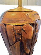 Two table lamps made from repurposed teak wood vases. The bases are tall and tapered, featuring a rugged, mosaic-like texture composed of various pieces of dark, polished teak wood with visible natural grain and crevices. Each lamp is topped with a large, warm-toned conical shade that is currently wrapped in protective clear plastic, emitting a soft, golden glow. The lamps are positioned on a clean white surface against a neutral background.
