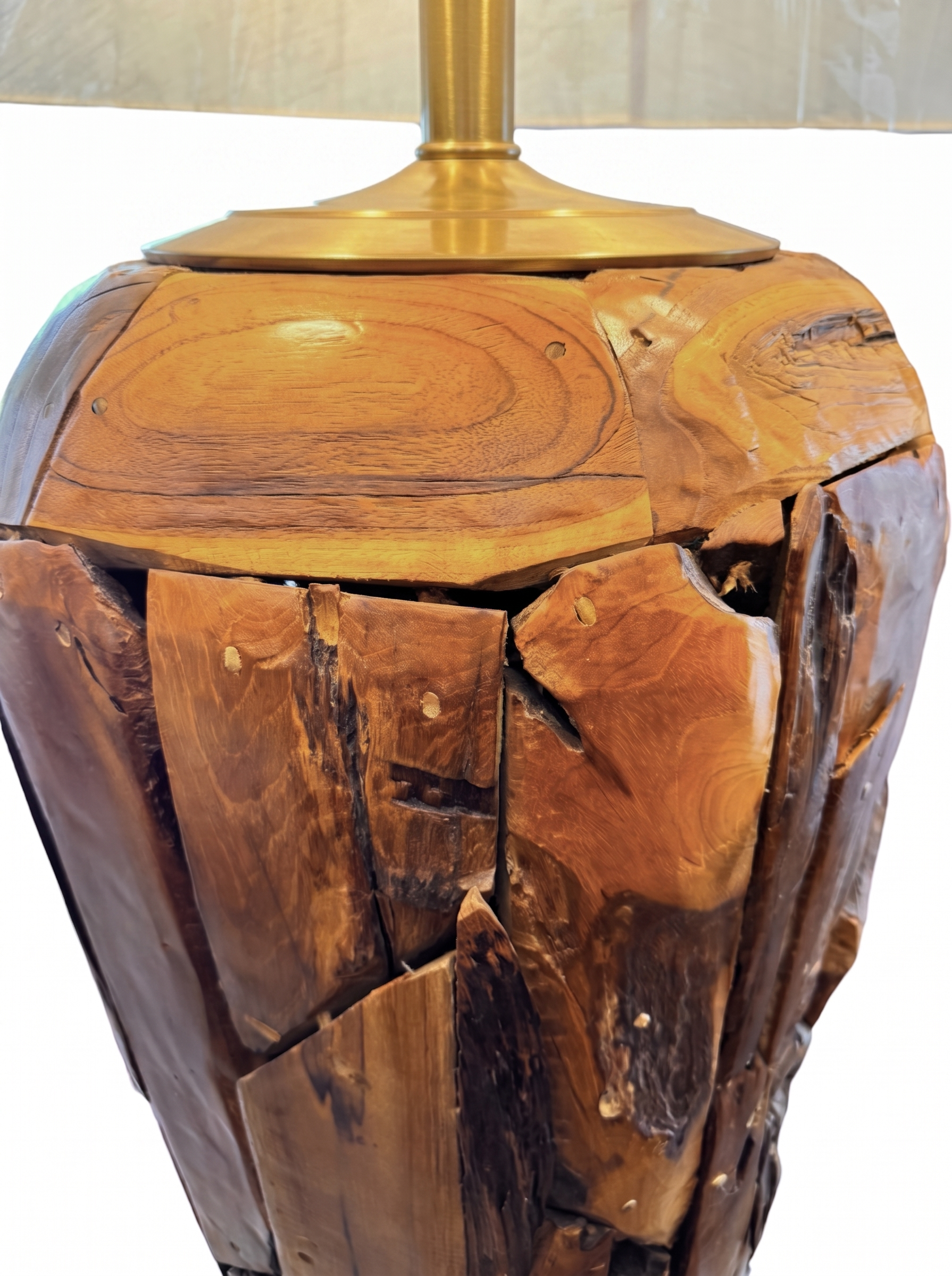 Two table lamps made from repurposed teak wood vases. The bases are tall and tapered, featuring a rugged, mosaic-like texture composed of various pieces of dark, polished teak wood with visible natural grain and crevices. Each lamp is topped with a large, warm-toned conical shade that is currently wrapped in protective clear plastic, emitting a soft, golden glow. The lamps are positioned on a clean white surface against a neutral background.
