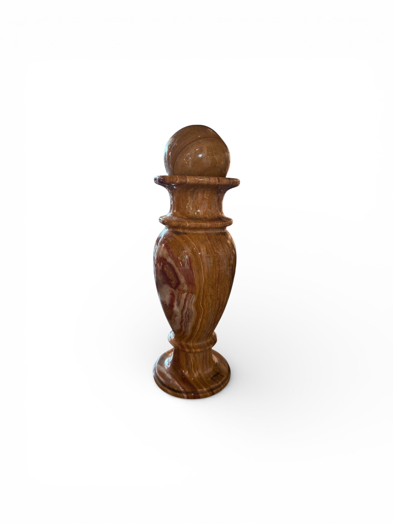 An elegant, hand-carved decorative vase made of polished Pakistani brown onyx. The vase features a classic, rounded urn shape with a tiered, flared neck and a solid matching spherical stone stopper resting on top. The stone displays rich, natural horizontal banding and swirling patterns in shades of honey, deep amber, rust-red, and cream. It stands on a small, integrated pedestal base against a clean white background, reflecting a high-gloss, mirror-like finish.