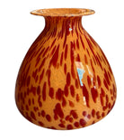 A hand-blown Italian Murano glass vase featuring a wide, bulbous body and a short, flared rim. The glass is a vibrant, translucent amber-orange base decorated with an organic "macchia" or spotted pattern of deep ruby red blotches. The red spots are elongated and vertical near the neck, becoming larger and more rounded toward the base. The surface has a high-gloss, reflective finish, showcasing mid-century modern craftsmanship.

