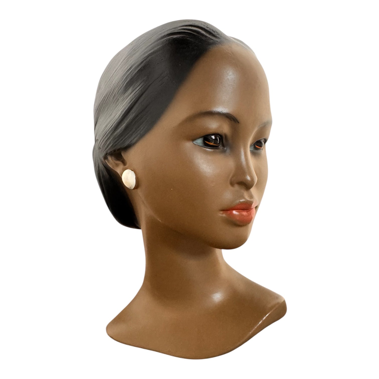 a vintage ceramic bust featuring a woman with brown skin, dark eyes, and orange lips. Her black hair is sleek and parted on the side, pulled back into a low style. She wears a small, round white earring in her right ear lobe. The bust includes her head, neck, and the upper part of her shoulders
