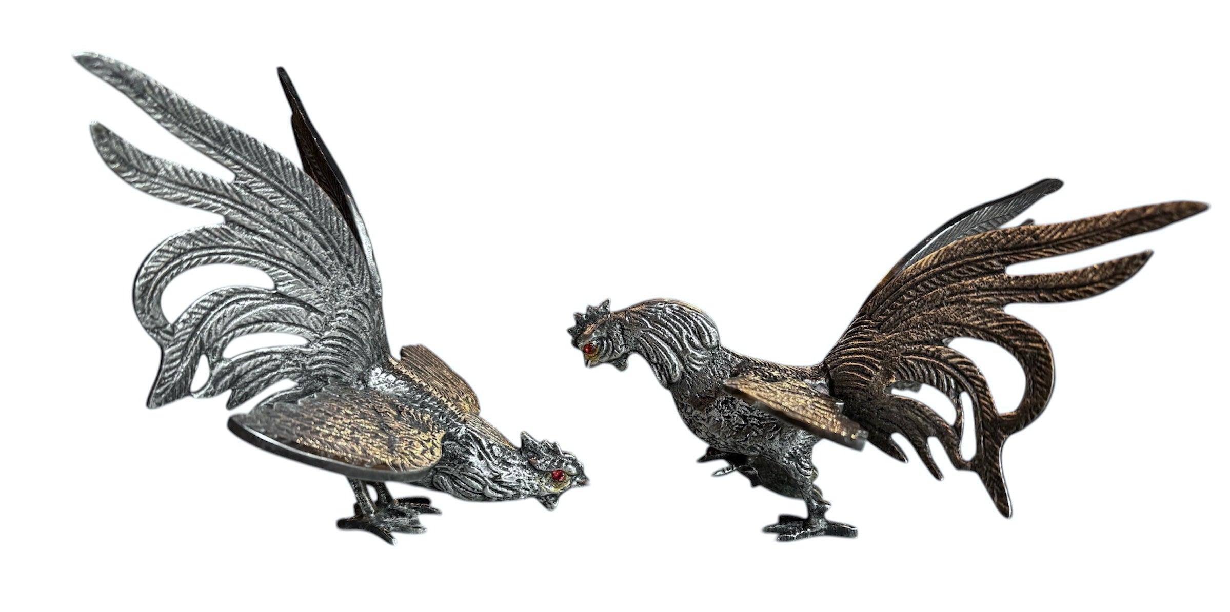 A pair of vintage, silver-plated brass fighting rooster figurines positioned in a confrontational stance against a white background. The rooster on the left is crouched low with its head down and tail feathers fanned high, while the rooster on the right stands more upright with its wings slightly flared. Both feature intricate feather detailing, a metallic patina showing hints of brass through the silver, and small, bright red glass eyes.