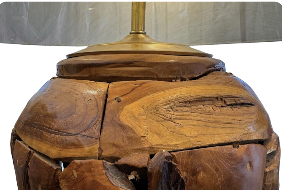 Two table lamps made from repurposed teak wood vases. The bases are tall and tapered, featuring a rugged, mosaic-like texture composed of various pieces of dark, polished teak wood with visible natural grain and crevices. Each lamp is topped with a large, warm-toned conical shade that is currently wrapped in protective clear plastic, emitting a soft, golden glow. The lamps are positioned on a clean white surface against a neutral background.
