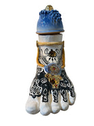 Handcrafted ceramic sculpture in the shape of a foot, decorated with intricate black and white patterns, 24KT gold accents, and topped with a blue lid featuring a gilt fish and head emerging from stylized waves.