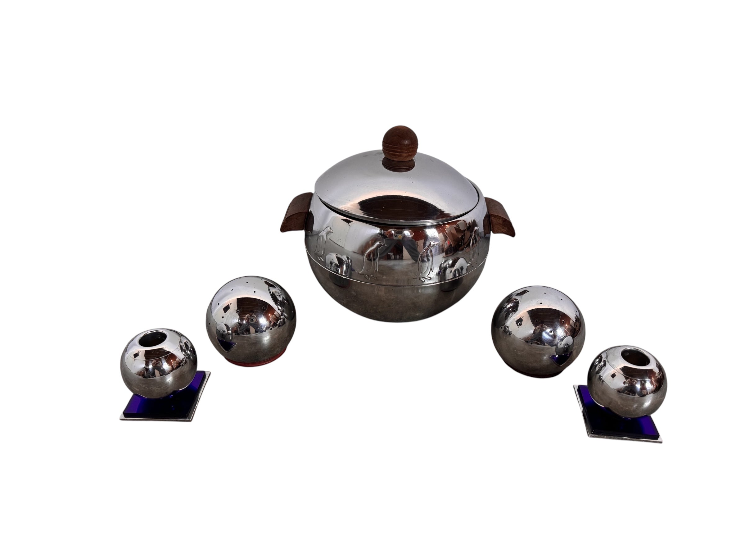 A set of vintage chrome-plated holloware featuring a central covered server with a relief penguin band and wood handles, flanked by two pairs of spherical shakers and candle holders on blue glass bases.