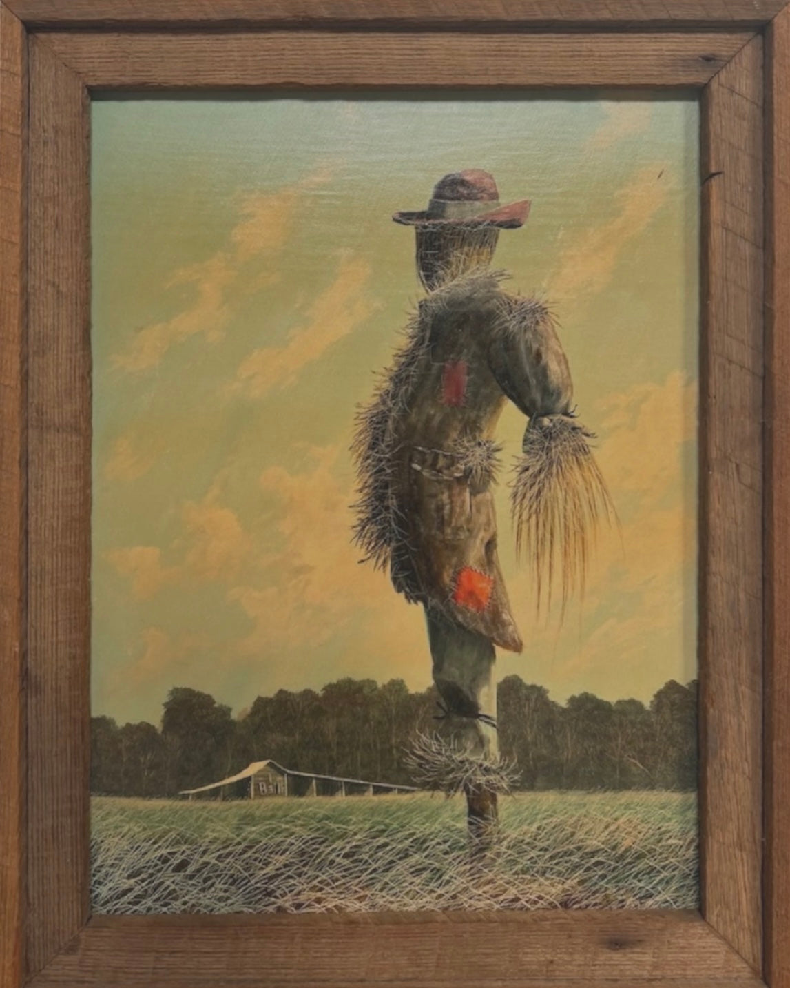 An oil painting on a wooden panel depicts a solitary scarecrow standing in a vast, grassy field under a pale yellow and blue sky. The scarecrow is shown from a three-quarters back view, wearing a brimmed hat and a tattered, patched brown coat with frayed straw bursting from its sleeves and edges. In the far left background, a long, white rustic barn sits before a dense line of green trees. The entire piece is enclosed in a thick, weathered, natural wood frame.
