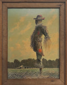 An oil painting on a wooden panel depicts a solitary scarecrow standing in a vast, grassy field under a pale yellow and blue sky. The scarecrow is shown from a three-quarters back view, wearing a brimmed hat and a tattered, patched brown coat with frayed straw bursting from its sleeves and edges. In the far left background, a long, white rustic barn sits before a dense line of green trees. The entire piece is enclosed in a thick, weathered, natural wood frame.