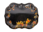 A vintage rectangular toleware serving tray with a scalloped, wavy edge and a black lacquered finish. The tray features hand-painted floral motifs in a traditional folk-art style. At the bottom center is a large, vibrant cluster of yellow and orange roses and daisies with green leaves. Smaller floral sprays and delicate gold filigree scrollwork decorate the corners and the upper rim. The center of the tray is open black space, showing light surface wear and minor scuffing consistent with age.