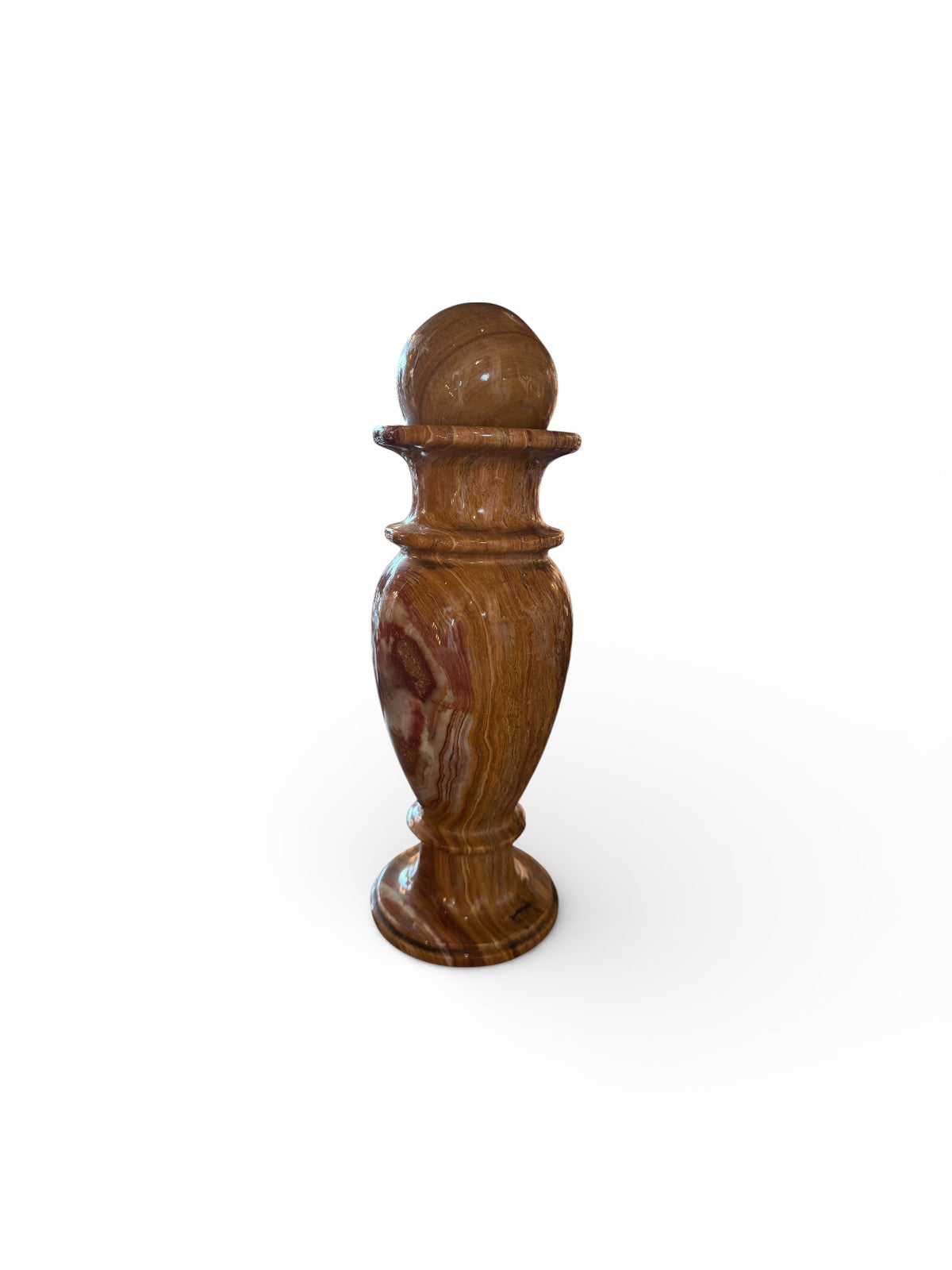 An elegant, hand-carved decorative vase made of polished Pakistani brown onyx. The vase features a classic, rounded urn shape with a tiered, flared neck and a solid matching spherical stone stopper resting on top. The stone displays rich, natural horizontal banding and swirling patterns in shades of honey, deep amber, rust-red, and cream. It stands on a small, integrated pedestal base against a clean white background, reflecting a high-gloss, mirror-like finish.